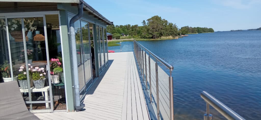 Ett modernt hus vid sjön med stora fönster och krukväxter ligger bredvid ett trädäck med utsikt över lugnt blått vatten och gröna träd en klar dag.
