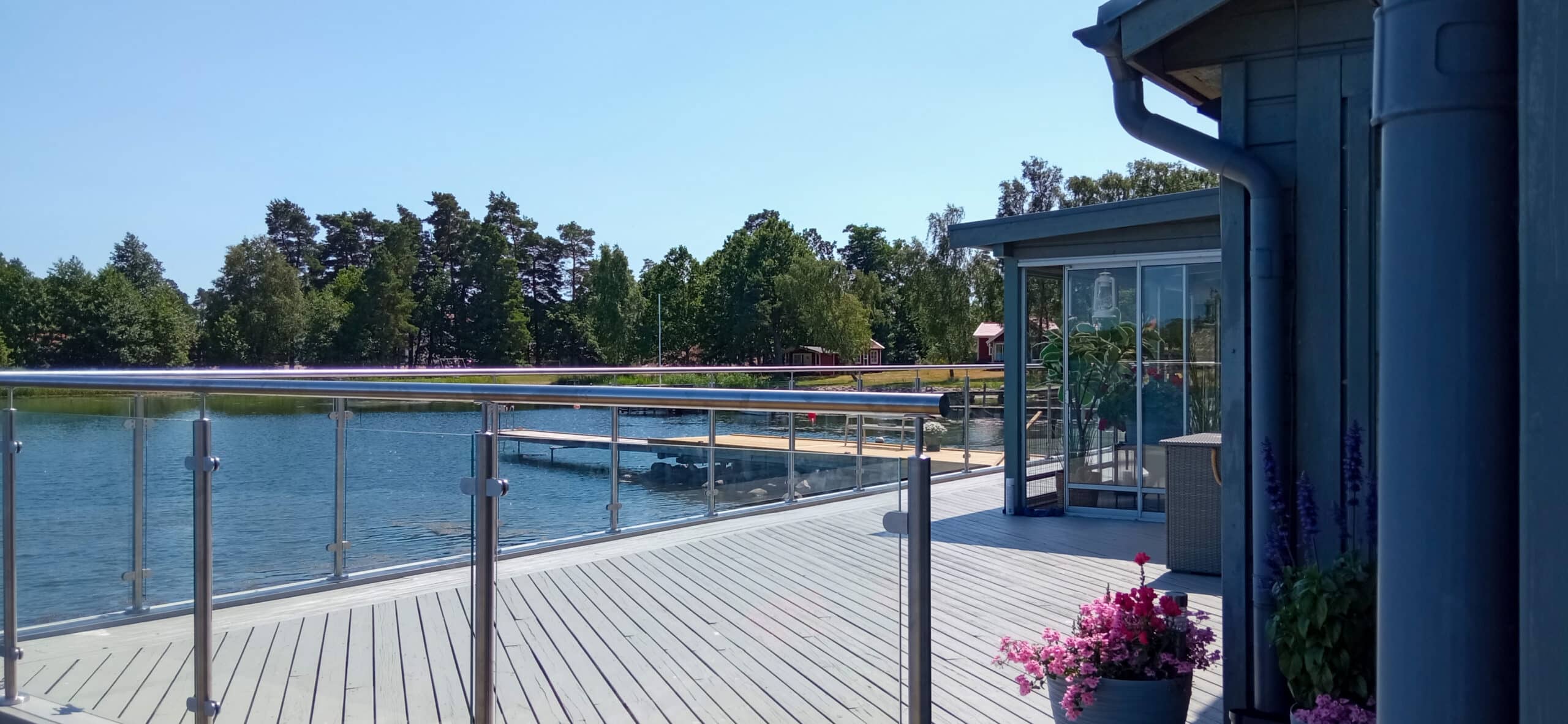 Ett trädäck med glasräcke har utsikt över ett lugnt vatten och träd; krukväxter sitter nära en blå byggnad under en klar himmel.