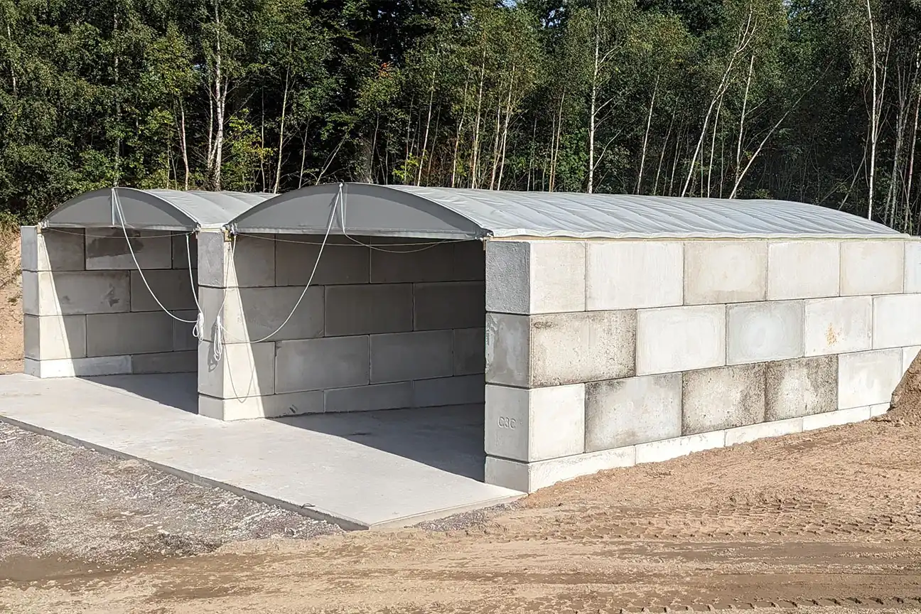 Två förråd med välvda tygtak och stora betongblock, byggda på en betongplatta nära ett skogsområde, ger ett hållbart skydd som liknar pooltäckning.