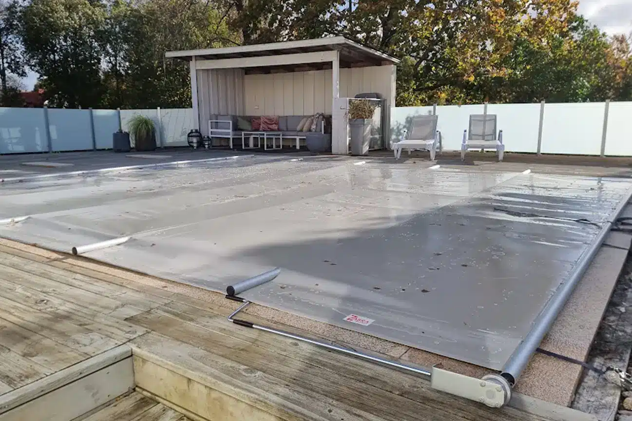 En täckt utomhuspool med ett transparent poolskydd och eleganta glasräcken, omgiven av ett trädäck, solstolar och en skuggig sittgrupp i bakgrunden.