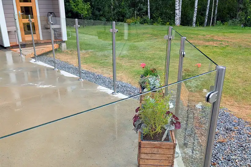Ett räcke i glas och metall omgärdar en uteplats i betong intill ett hus, med blomkrukor och grönska längs kanten; träd och gräsmatta i bakgrunden.