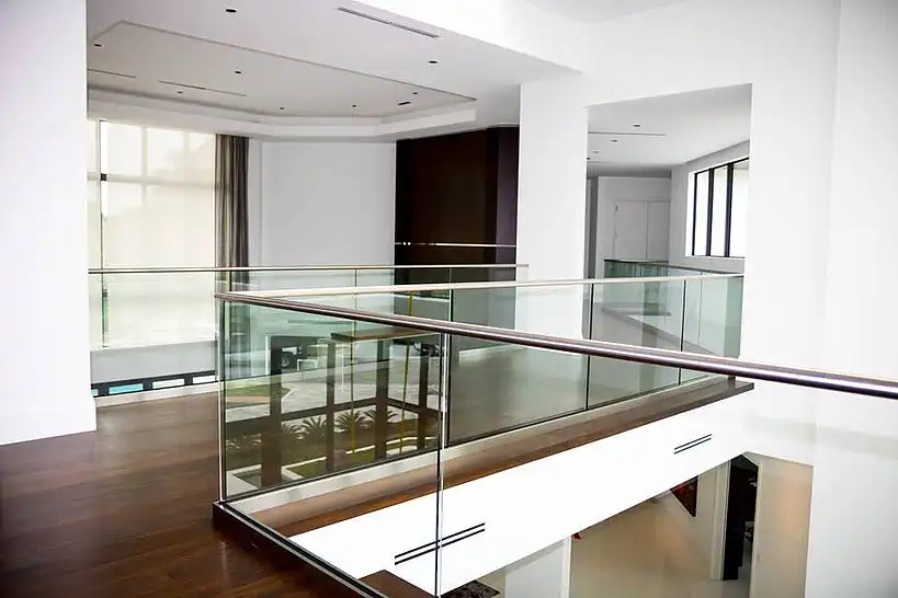 Modern inomhusbalkong med glasräcken, mörka trägolv och vita väggar, med utsikt över ett nedre plan med stora fönster och naturligt ljus.