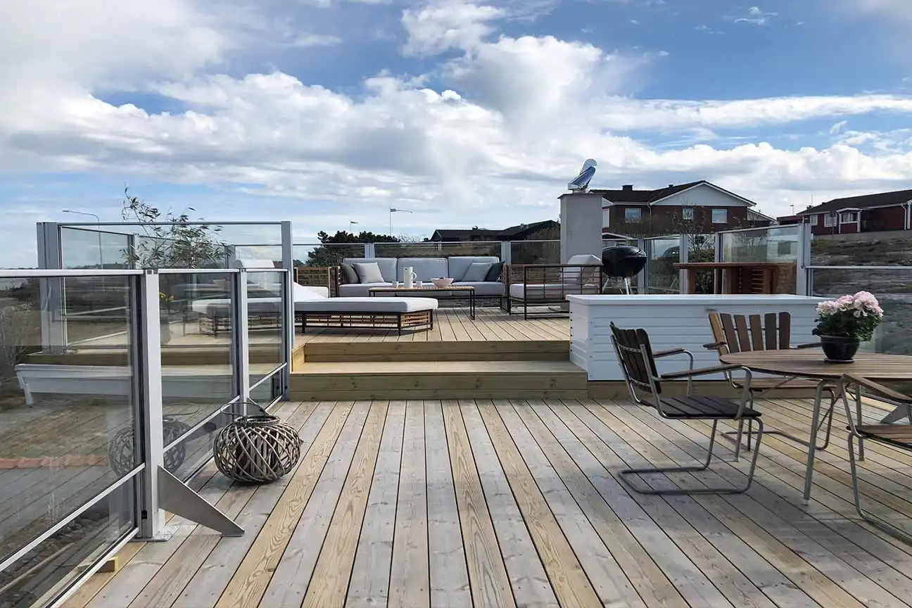 Ett modernt takterrass i trä med Glasräcken, uteservering, matbord med stolar, krukväxter och en delvis molnig himmel ovanför.
