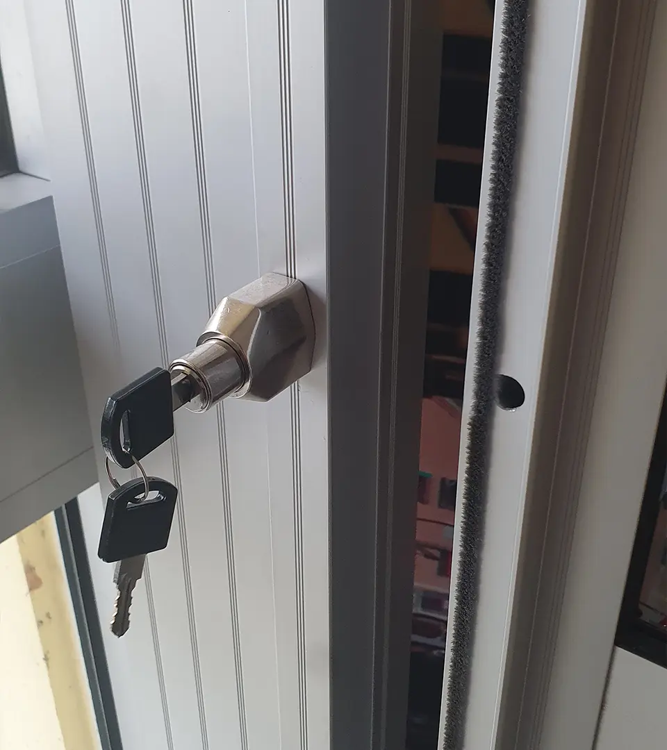 nyckel1 Närbild på ett dörrlås av metall med två nycklar i. Dörren står på glänt, vilket avslöjar låsmekanismen och tätningslisterna.