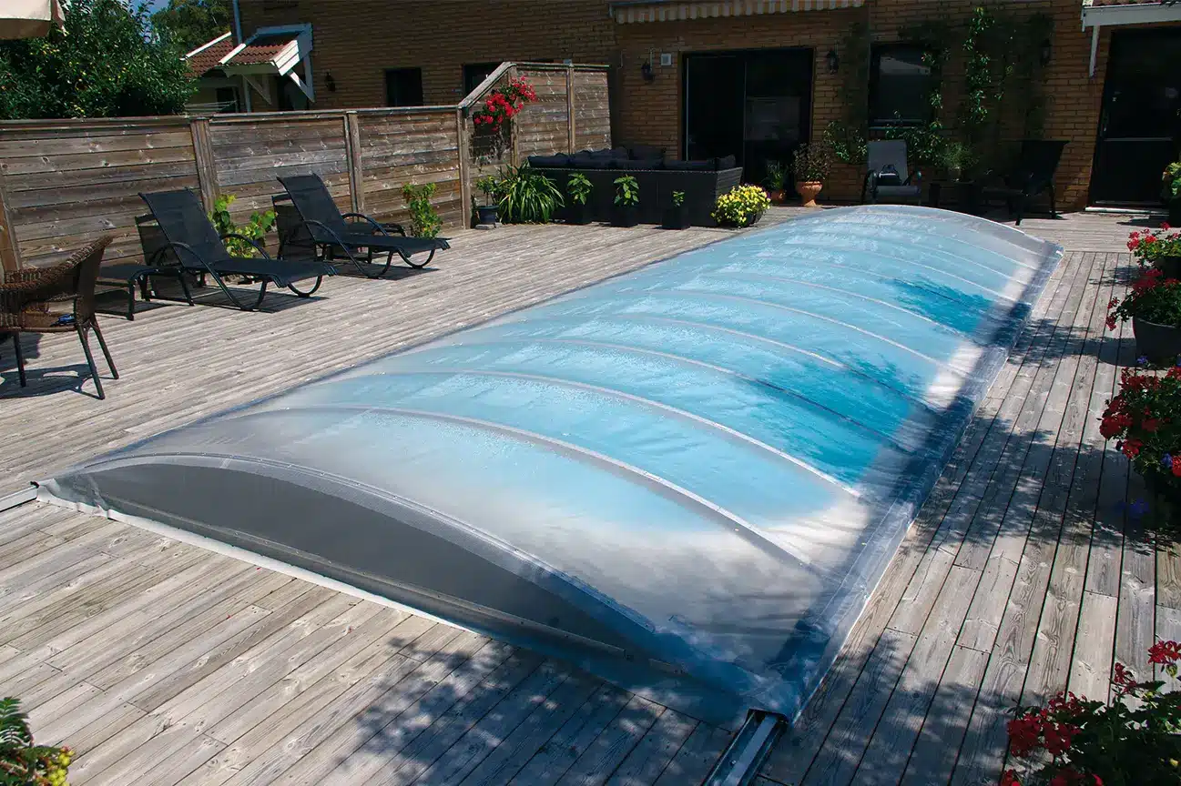 En rektangulär pool med ett transparent, svängt lock ligger på ett trädäck med glasräcken, solstolar, krukväxter och ett insynsskyddande staket.