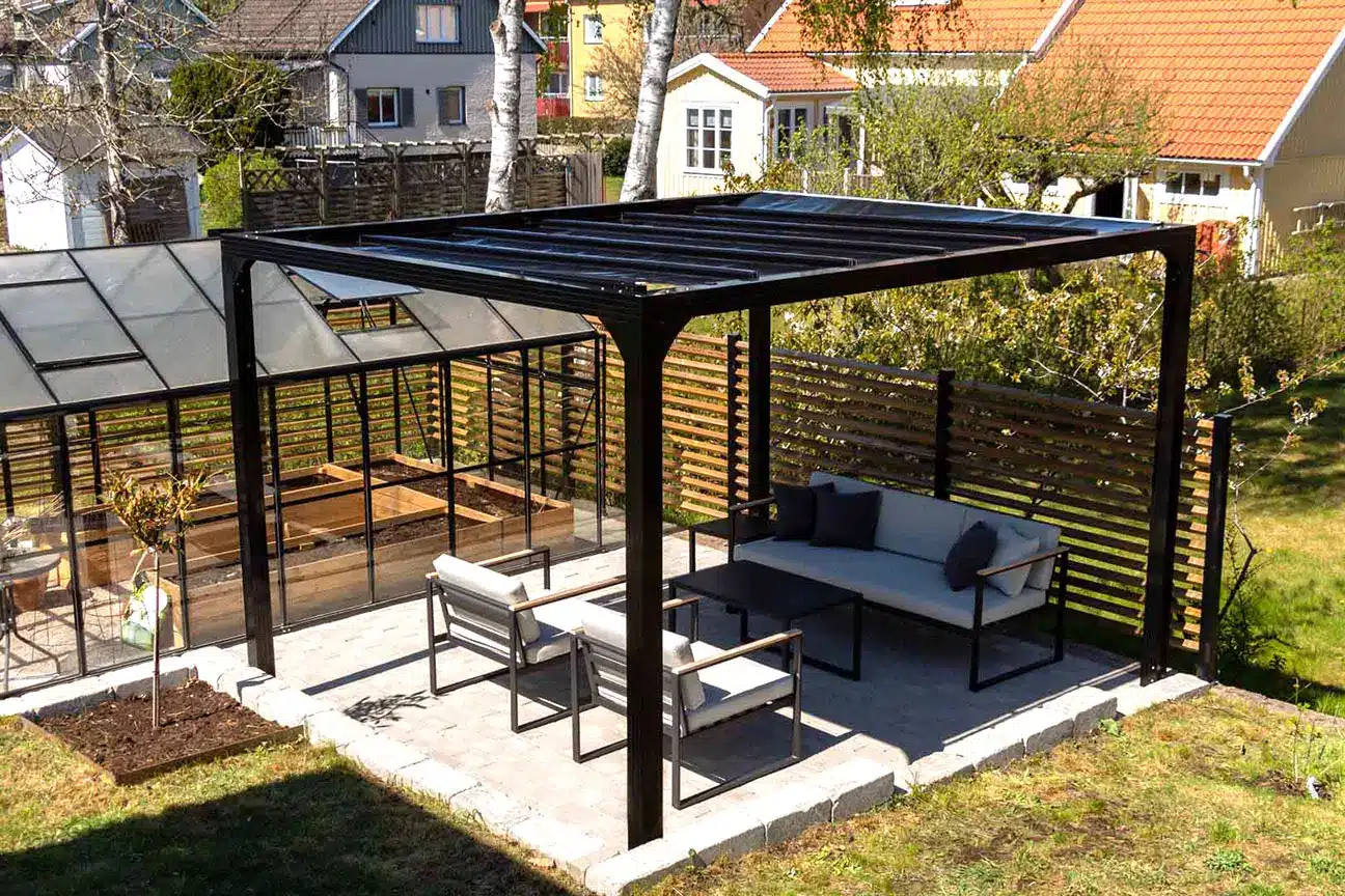 svart pergola står i en trädgård med stensättning under och vita utemöbler