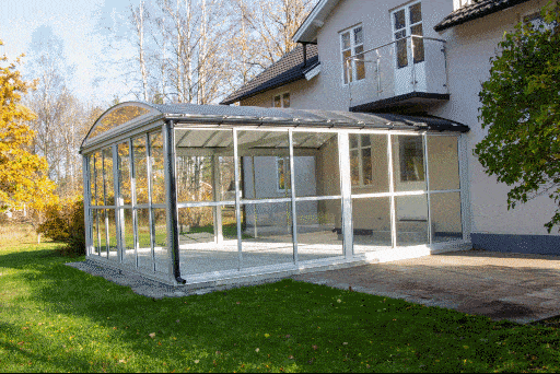 En inglasad uteplats med ett böjt svart tak står i en trädgård med grönt gräs, träd och närliggande byggnader synliga.