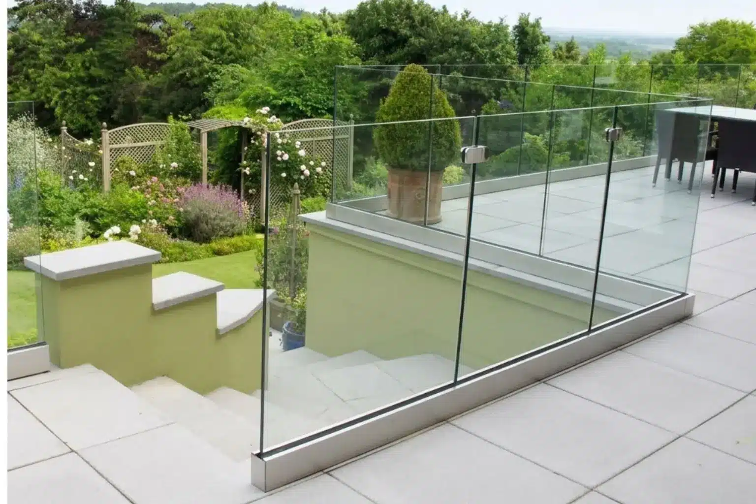Ett modernt uterum med en terrass i grå stenplattor och ett glasräcke runt en nedgång till en trädgård. Nedanför syns en grönskande trädgård med blommande buskar, spaljéer och träd. Uppe på terrassen står en stor kruka med en buske samt utemöbler i bakgrunden.
