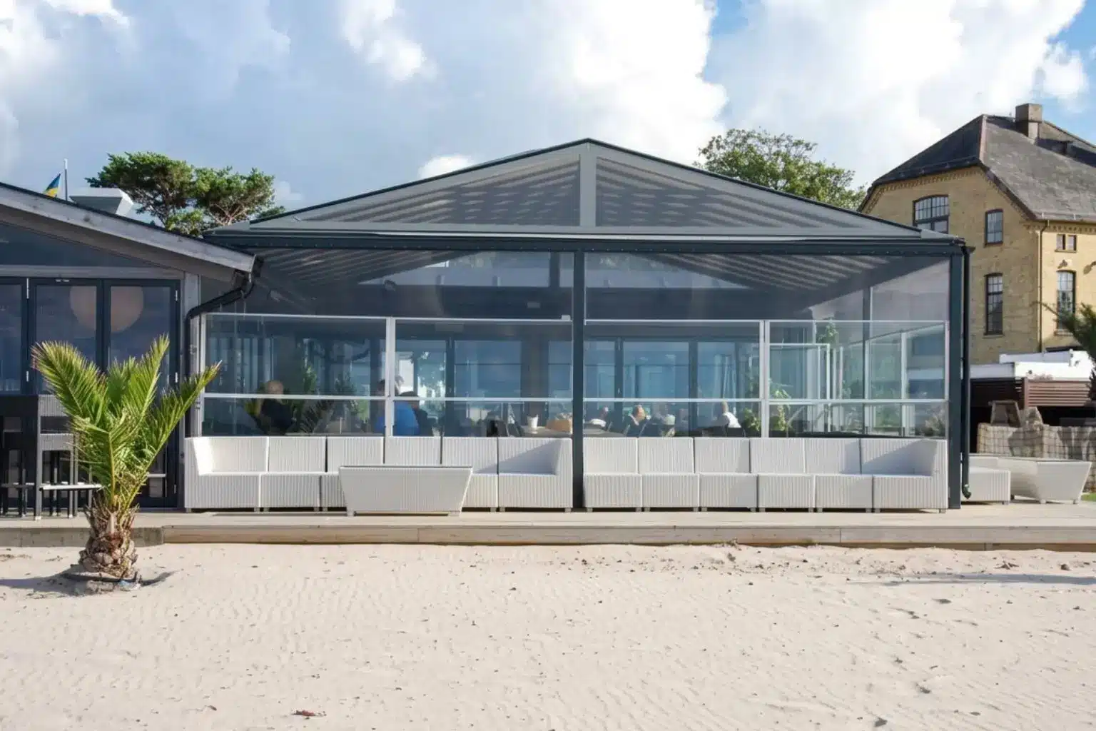 En strandnära uteservering med ett modernt glastak och glasräcke. Framför byggnaden syns sand och en liten palm, och inuti finns vita soffor och sittplatser där gäster sitter. I bakgrunden syns en byggnad och blå himmel med moln.